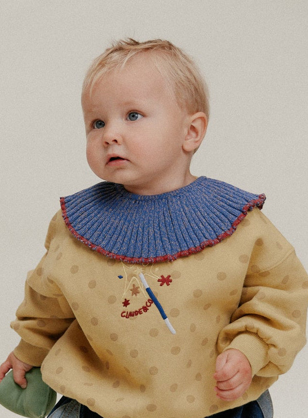 A baby standing and wearing a mustard yellow knit sweater with a blue ribbed collar, blue patchwork pants, and white shoes, photographed against a plain light beige studio background.