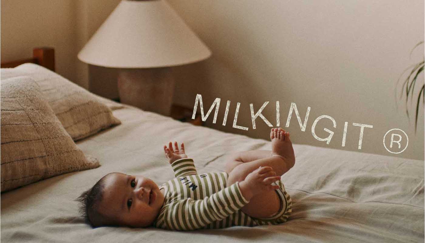 A baby wearing a green and white striped long-sleeve bodysuit lies on their back on a bed in a softly lit bedroom. The brand name 'MILKING IT' is displayed in white text over the scene.